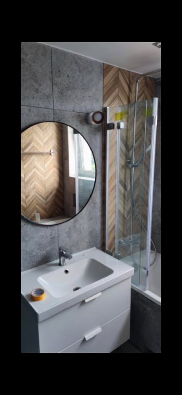 Wykończona łazienka z umywalką na szafce, okrągłym lustrem i kabiną prysznicową. Ściany wyłożone płytkami imitującymi beton i drewnem ułożonym w jodełkę.