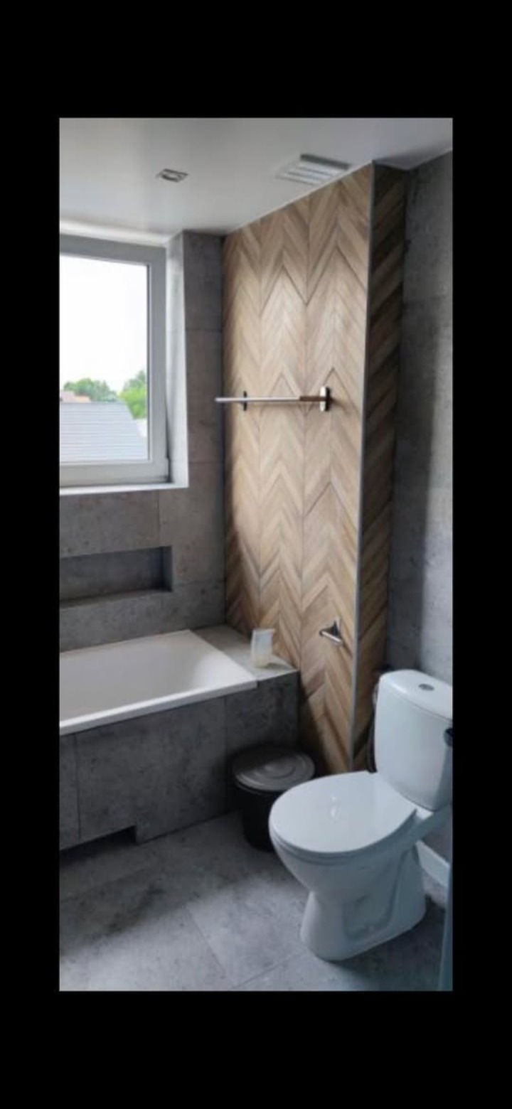 Nowoczesna łazienka z wanną w zabudowie z szarego betonu i toaletą. Na ścianie dekoracyjna mozaika w jodełkę. Okno z widokiem na zieleń.