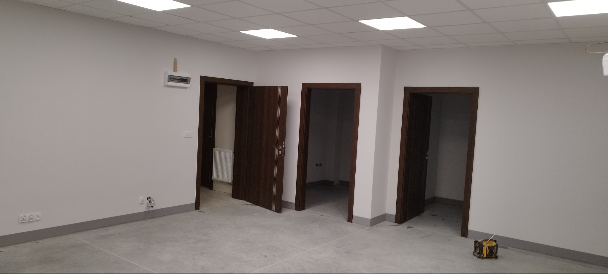 Wnętrze po remoncie: trzy drewniane drzwi z ościeżnicami, świeżo pomalowane ściany w neutralnym odcieniu, płytki podłogowe i narzędzie pomiarowe w rogu.