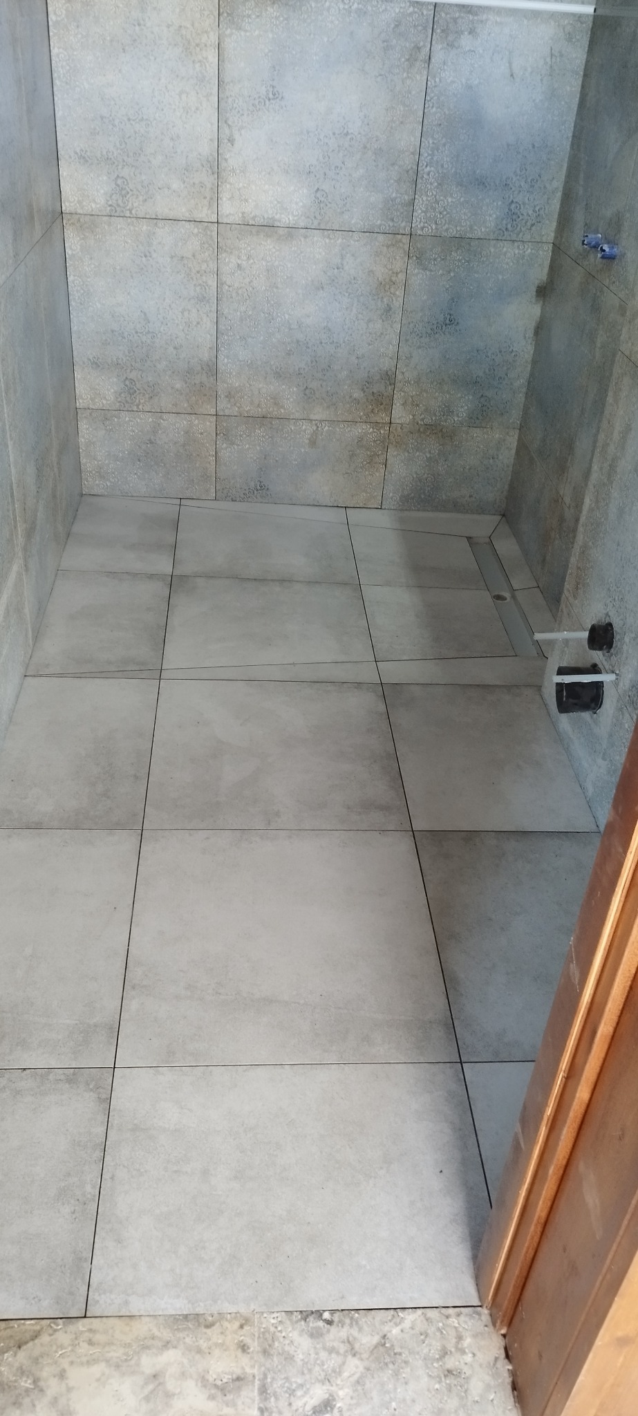 Wykończona kabina prysznicowa z szarymi, kwadratowymi płytkami na ścianach i podłodze. Widoczny odpływ liniowy i fragment drewnianej framugi drzwi.