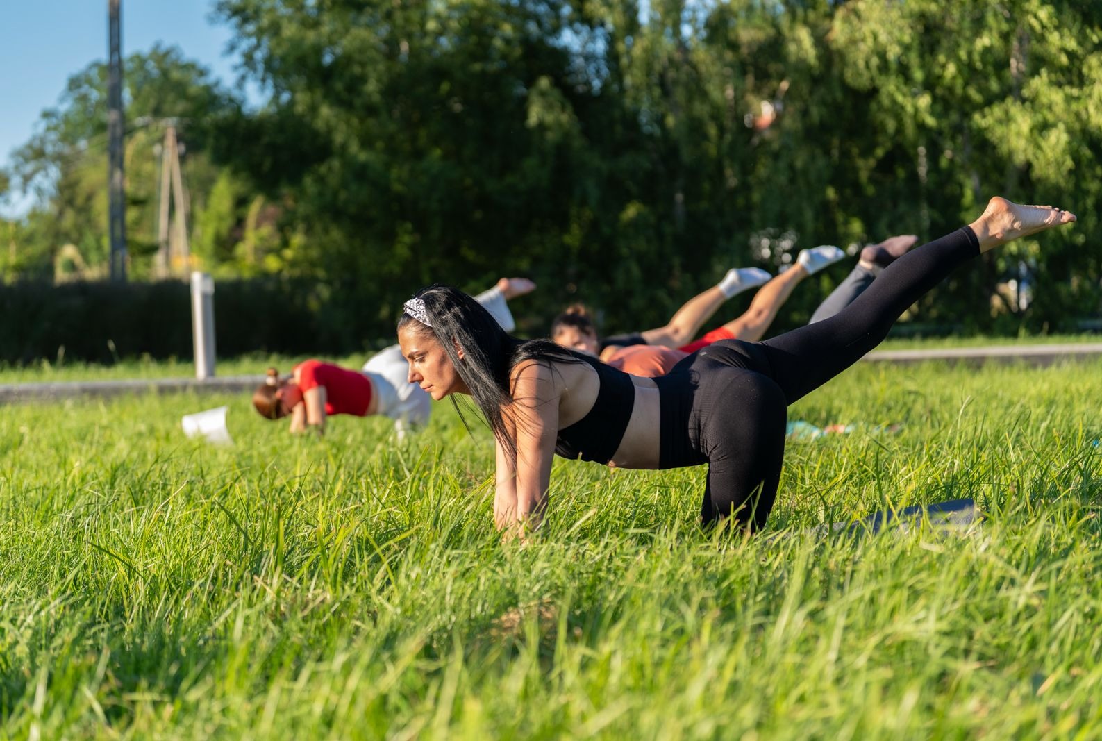 Grupa osób ćwiczy na trawie w parku, unosząc nogi do góry. Kobieta na pierwszym planie w czarnym stroju sportowym i opasce na włosach. Poranne słońce oświetla zieloną trawę.