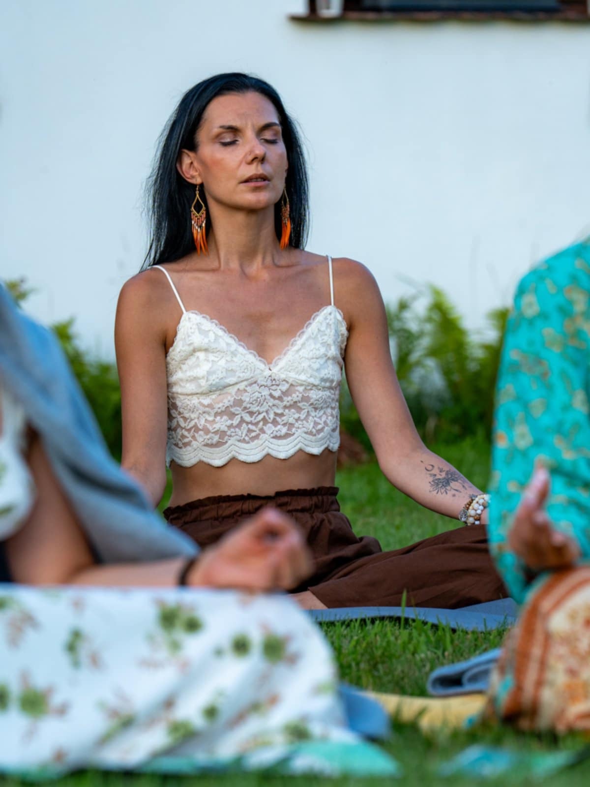 Kobieta w koronkowej bluzce i brązowych spodniach medytuje na trawie z innymi osobami, skupiona twarz, kolczyki z piór.