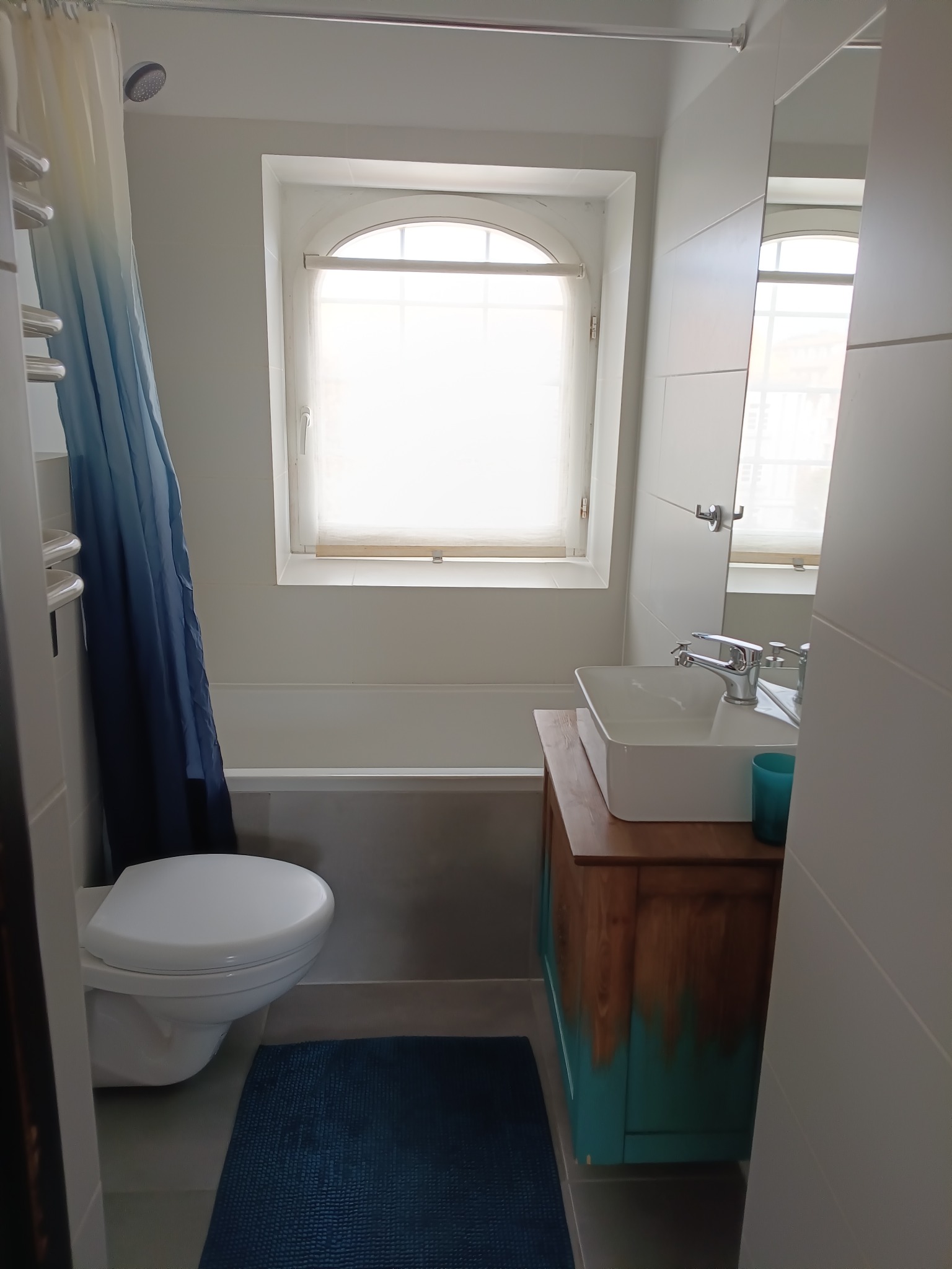 Jasna łazienka z wanną pod oknem, umywalką na drewnianej szafce z turkusowym akcentem i toaletą. Niebieski dywanik i zasłona prysznicowa dodają koloru.