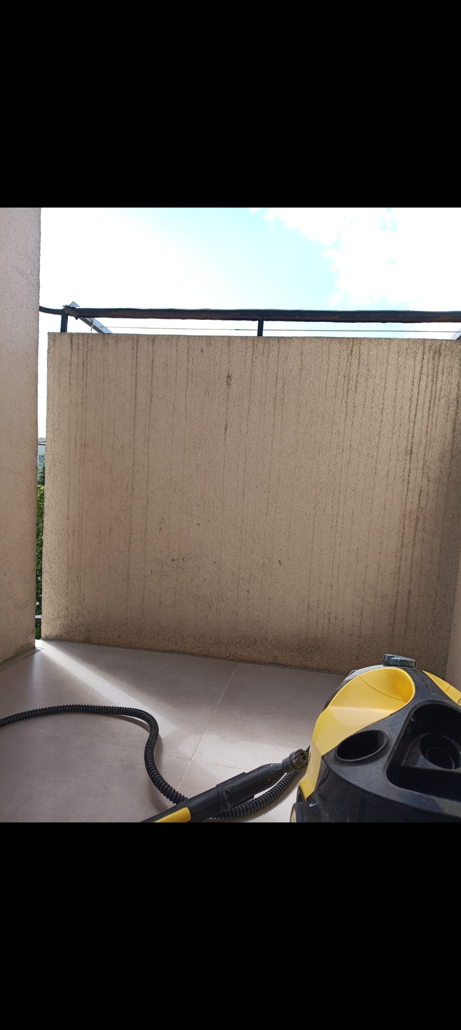 Balkon przed czyszczeniem parowym. Widoczne zabrudzenia na ścianie i podłodze. W rogu stoi żółto-czarny parownica z wężem. W tle fragment nieba i zieleń.