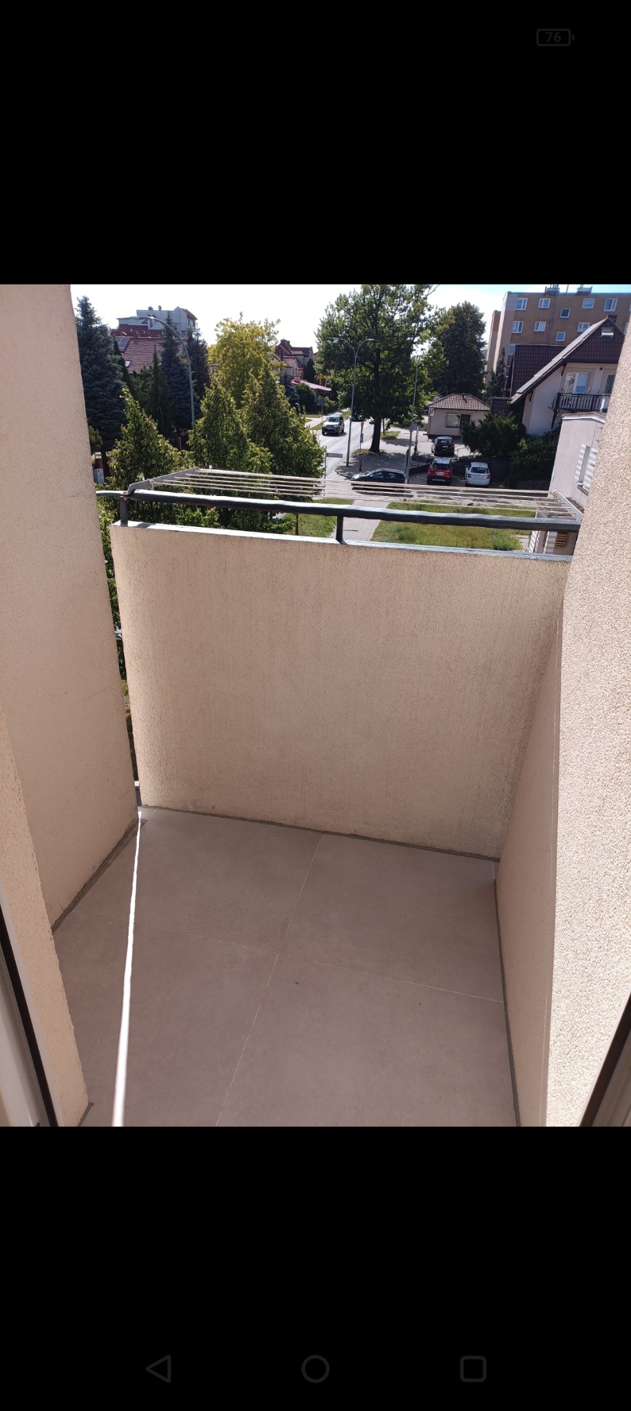 Jasny balkon z widokiem na ulicę i zieleń. Świeżo ułożone płytki podłogowe. Metalowa suszarka na pranie. Słoneczny dzień.