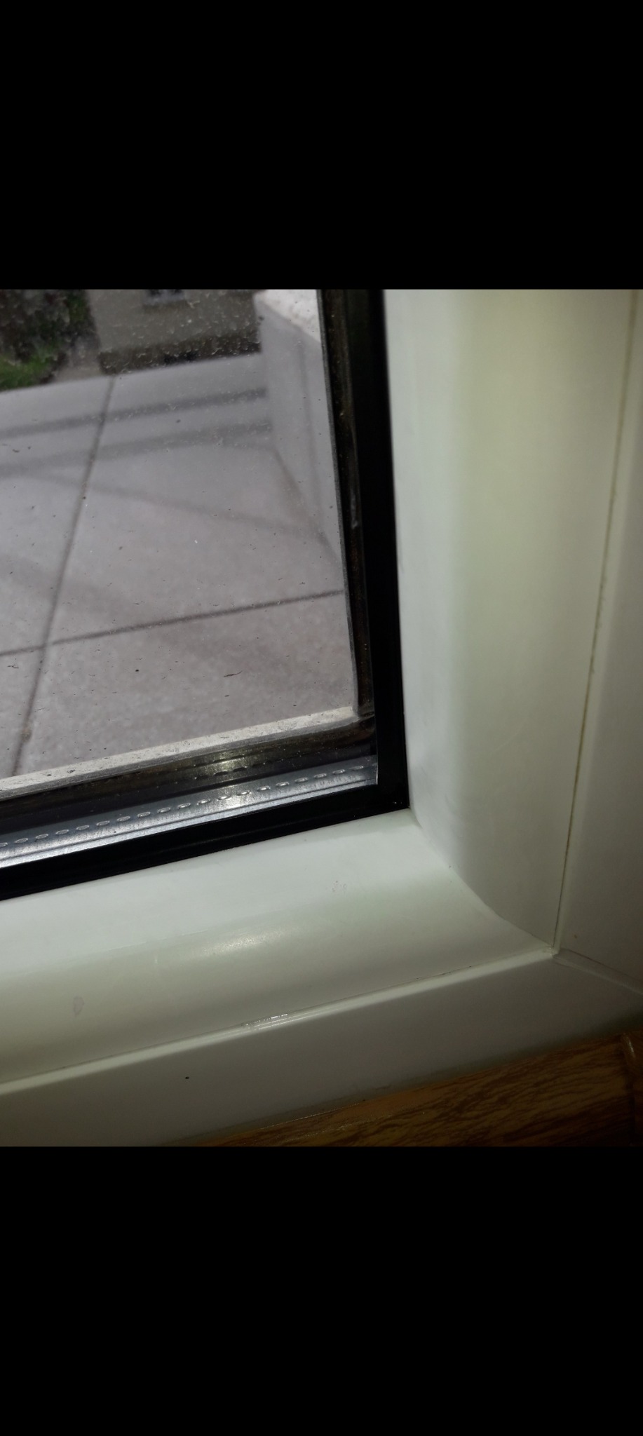 Fragment okna PCV z widocznymi zabrudzeniami na szybie i ramie. Ujęcie z bliska, ukazujące detale i fakturę materiałów. Widok na zewnątrz przez brudną szybę.