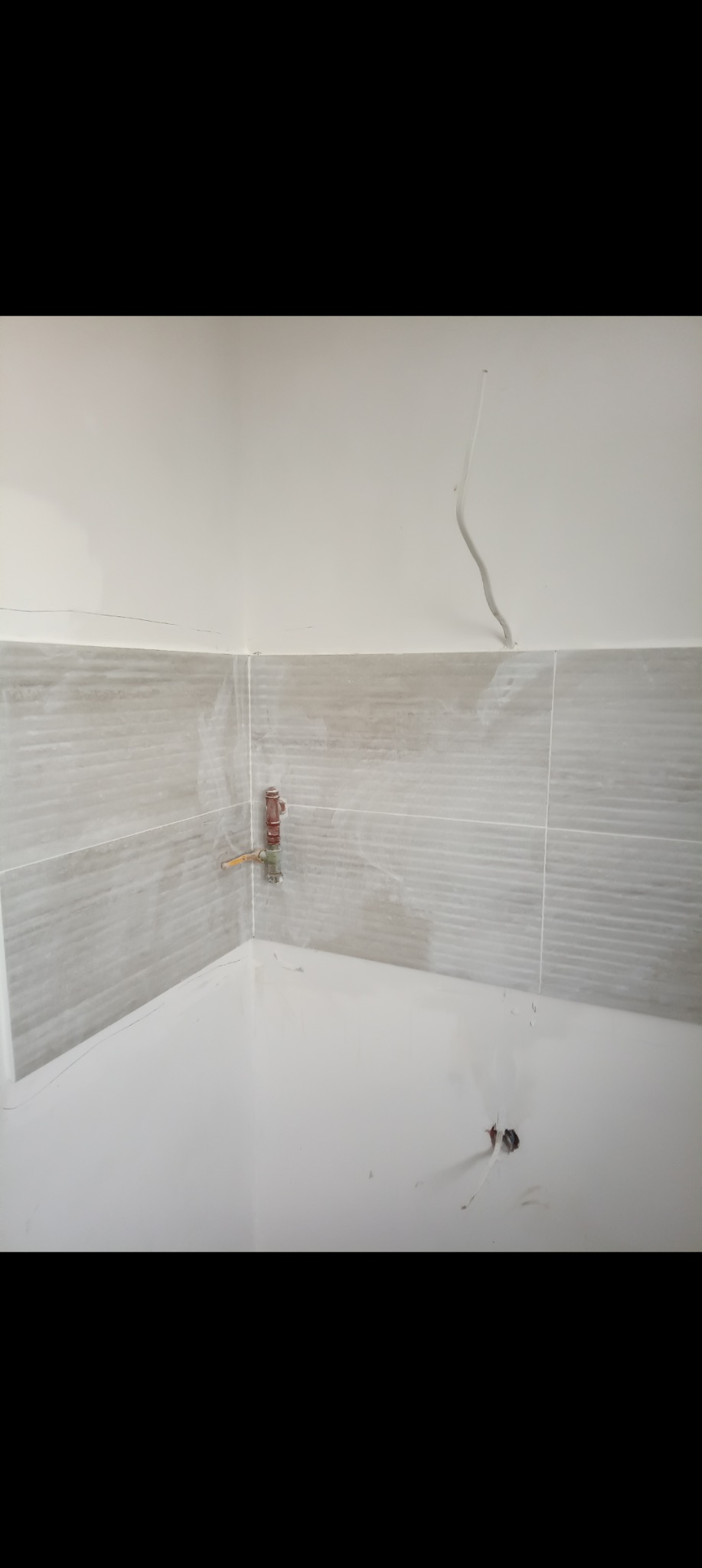 Fragment łazienki w trakcie remontu: szare płytki na ścianie, wystające rury i kable. Widoczne ślady po pracach wykończeniowych.