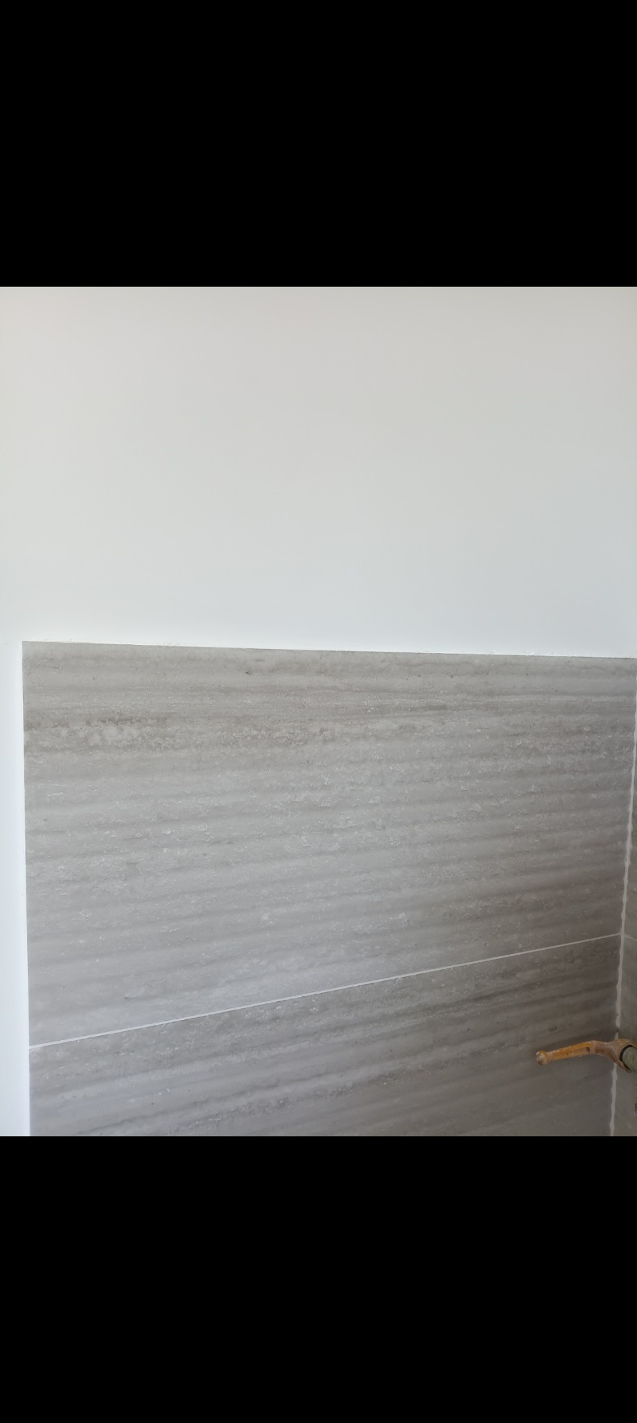 Ściana wyłożona szarymi, poziomymi płytkami imitującymi drewno, z widocznym fragmentem instalacji wodnej w prawym dolnym rogu. Minimalistyczne wykończenie wnętrza.