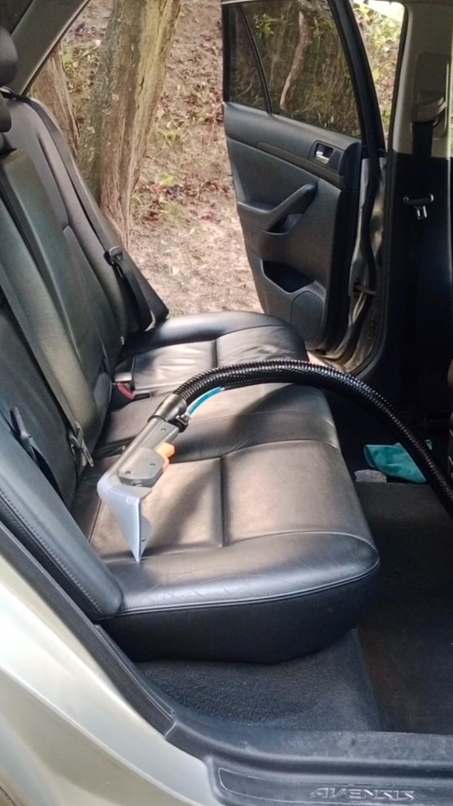 Czyszczenie skórzanej tapicerki samochodowej odkurzaczem piorącym. Widoczne otwarte drzwi auta i zieleń w tle. Detailing wnętrza pojazdu.