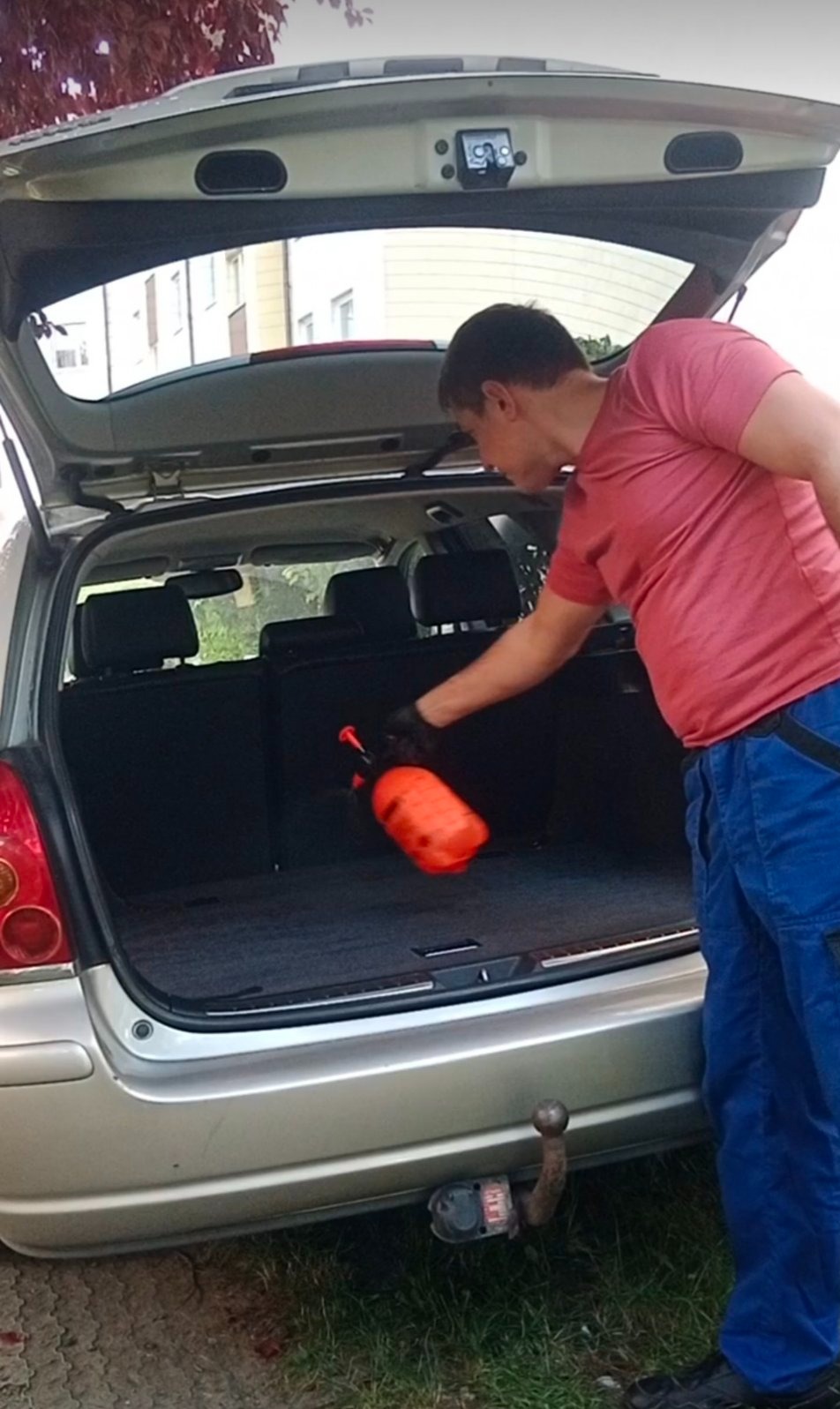 Mężczyzna w rękawicach spryskuje tapicerkę bagażnika kombi pomarańczowym spryskiwaczem ciśnieniowym. Otwarte drzwi bagażnika, widoczne tylne siedzenia.