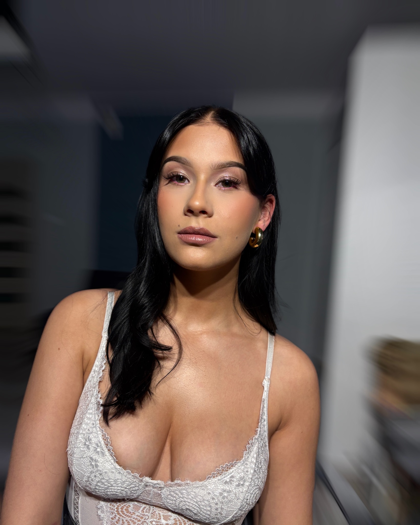 Portret kobiety z delikatnym makijażem, podkreślającym oczy i usta, w białej koronkowej bieliźnie i złotych kolczykach, na rozmytym tle.