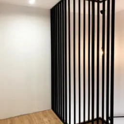 Nowoczesna, czarna balustrada z pionowych, metalowych prętów, tworząca ażurową ściankę działową w jasnym wnętrzu z drewnianą podłogą. Minimalistyczny design.