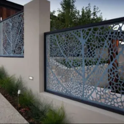 Dekoracyjny panel ogrodzeniowy z ażurowym wzorem drzewa, wkomponowany w mur z oświetleniem. Widoczne trawy ozdobne wzdłuż ścieżki.