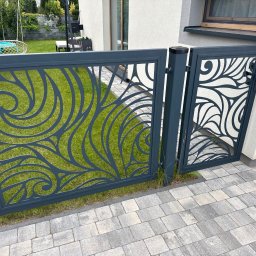 Lubner Schody - Nowoczesna, metalowa brama z ażurowym, roślinnym wzorem, pomalowana na grafitowo. Brama zamontowana przy jasnym budynku, widoczny trawnik i kostka brukowa.