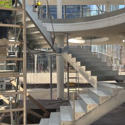 Lubner Schody - Montaż spiralnych schodów z metalu na zewnątrz budynku. Widoczny pracownik na rusztowaniu i elementy konstrukcji. Jasne światło dzienne.