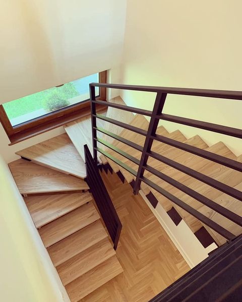 Drewniane schody z ciemną, minimalistyczną balustradą w nowoczesnym wnętrzu. Widok z góry, z okna wpada naturalne światło. Parkiet ułożony w jodełkę.