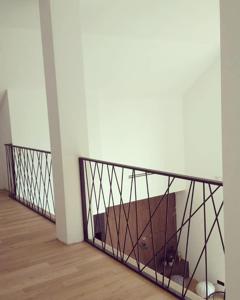 Nowoczesne balustrady z czarnych prętów tworzących geometryczny wzór, widoczne z góry. Jasne wnętrze, drewniana podłoga, białe ściany. Minimalistyczny design.