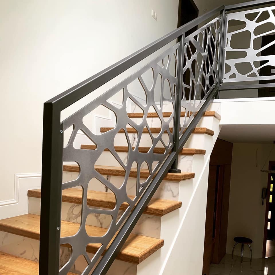 Nowoczesne schody z drewnianymi stopniami i balustradą z metalu o ażurowym wzorze. Białe ściany i marmurowe wykończenie dodają elegancji wnętrzu.