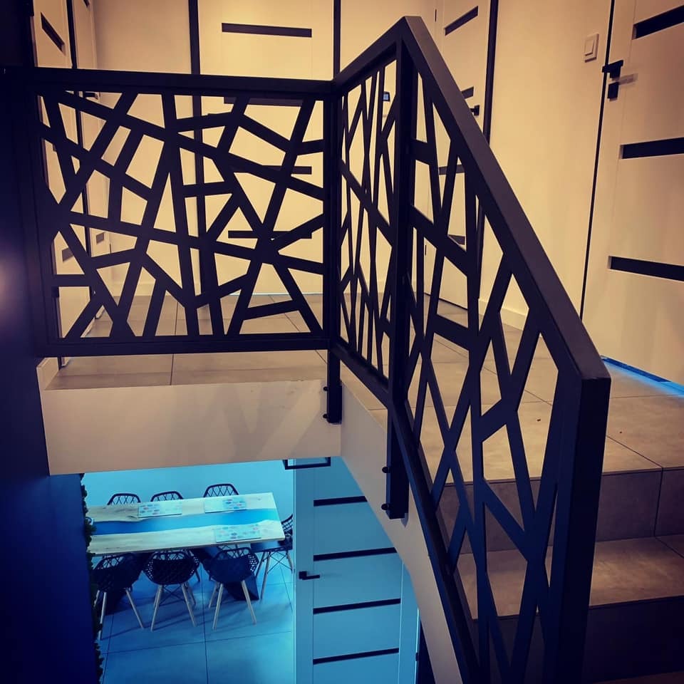 Nowoczesna, czarna balustrada schodowa z geometrycznym wzorem, widoczna z góry. W tle jasne ściany z dekoracyjnymi paskami i fragment pomieszczenia z stołem.