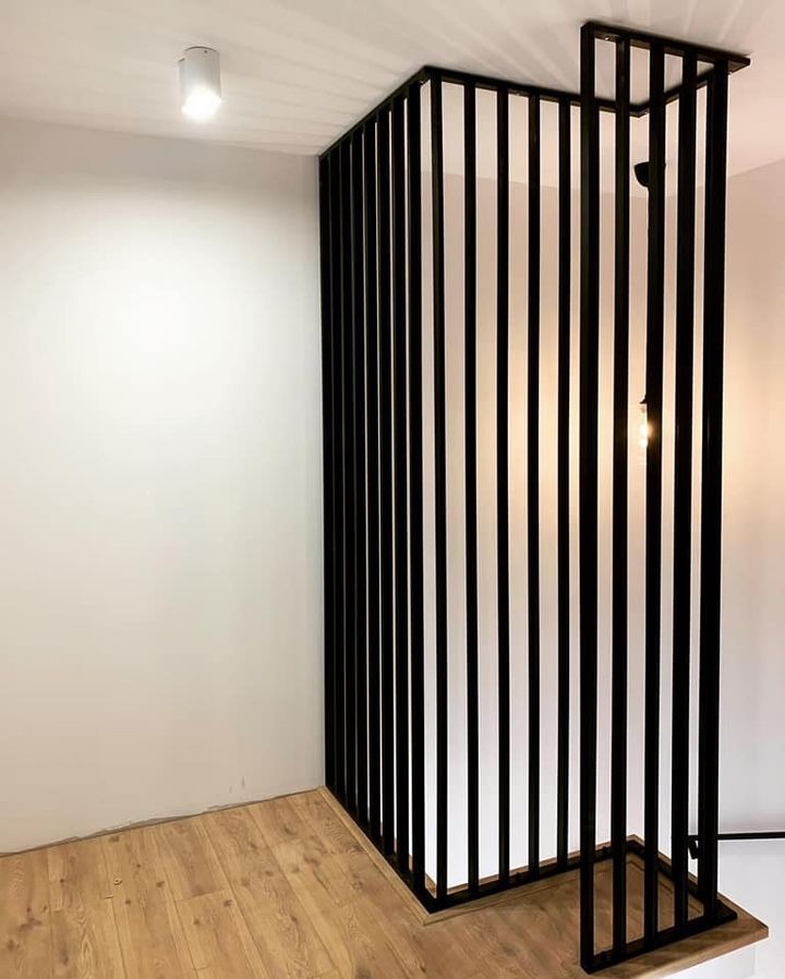 Nowoczesna, czarna balustrada z pionowych, metalowych prętów, tworząca ażurową ściankę działową w jasnym wnętrzu z drewnianą podłogą. Minimalistyczny design.