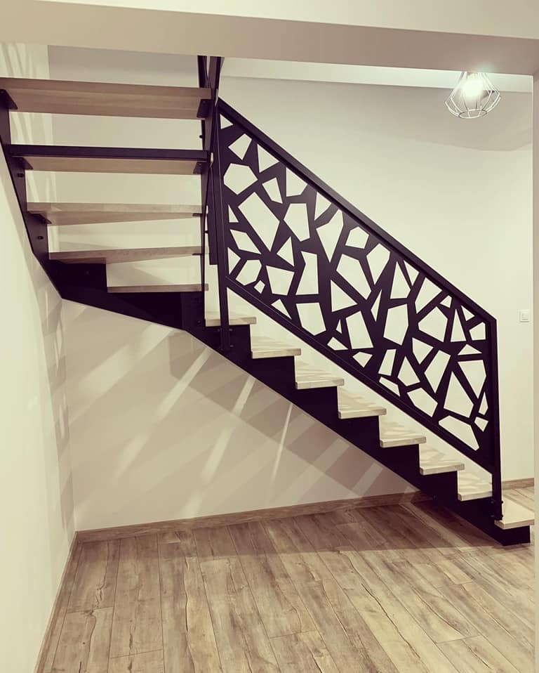 Nowoczesne schody z drewnianymi stopniami i czarną, ażurową balustradą o geometrycznym wzorze, widoczne z dołu, w jasnym wnętrzu.