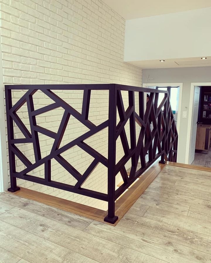 Nowoczesna balustrada w kolorze czarnym o geometrycznym wzorze na tle białej ściany z cegły. Balustrada zamontowana na drewnianej podłodze, widoczny fragment schodów.
