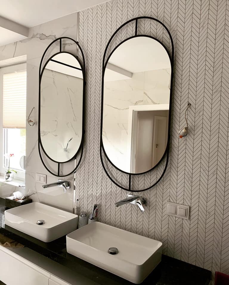 Dwa owalne lustra w czarnych, metalowych ramach nad umywalkami w łazience w Łomży. W tle wzorzysta tapeta i marmurowe wykończenia.
