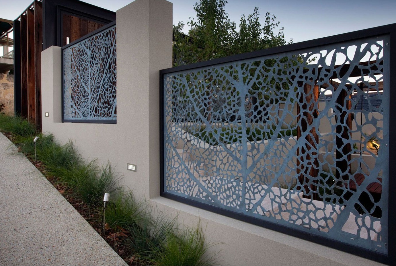 Dekoracyjny panel ogrodzeniowy z ażurowym wzorem drzewa, wkomponowany w mur z oświetleniem. Widoczne trawy ozdobne wzdłuż ścieżki.