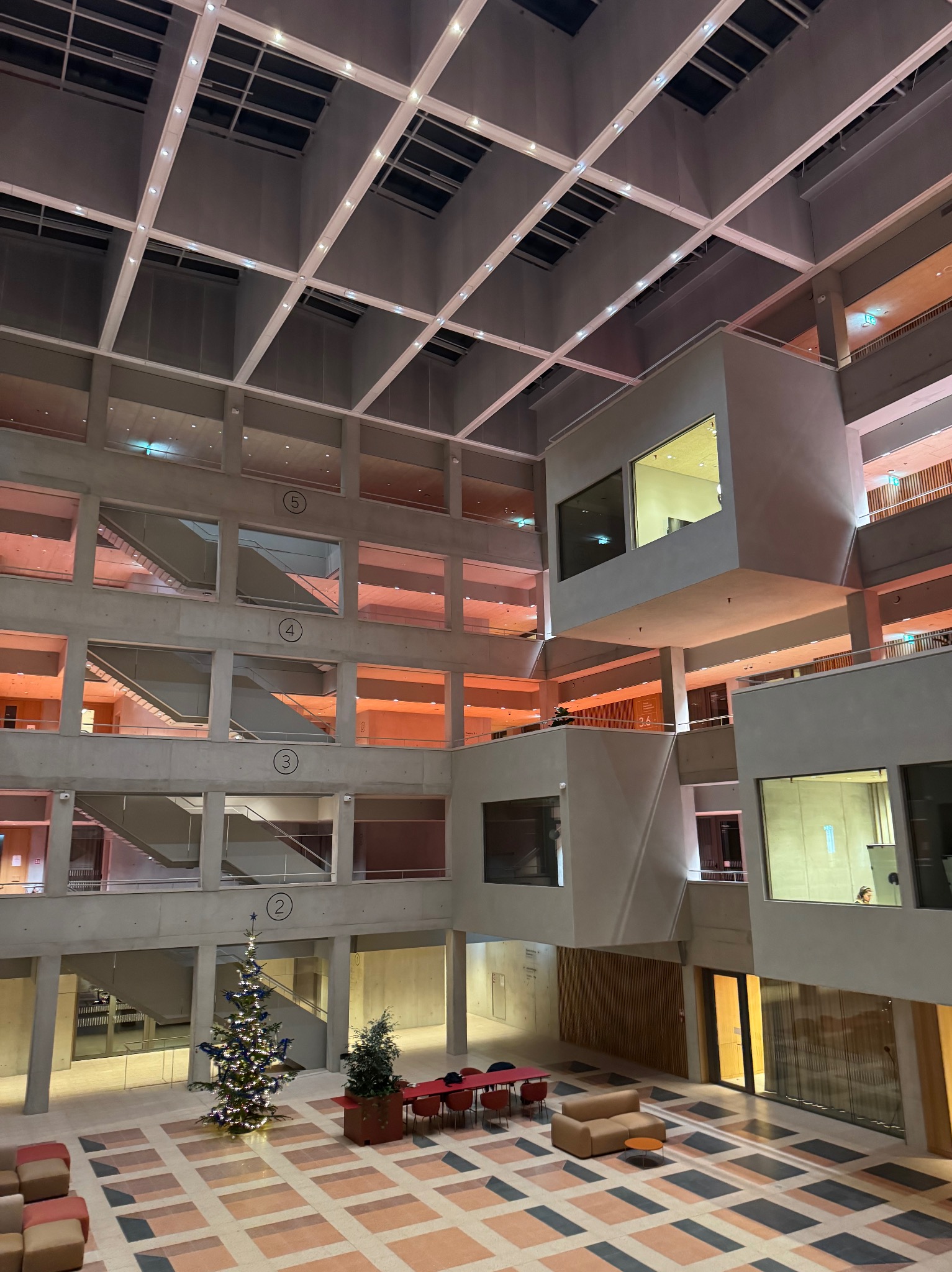 Wnętrze nowoczesnego budynku z betonowymi schodami i balustradami, widok z dołu na kilka pięter. W tle widoczne dekoracje świąteczne.