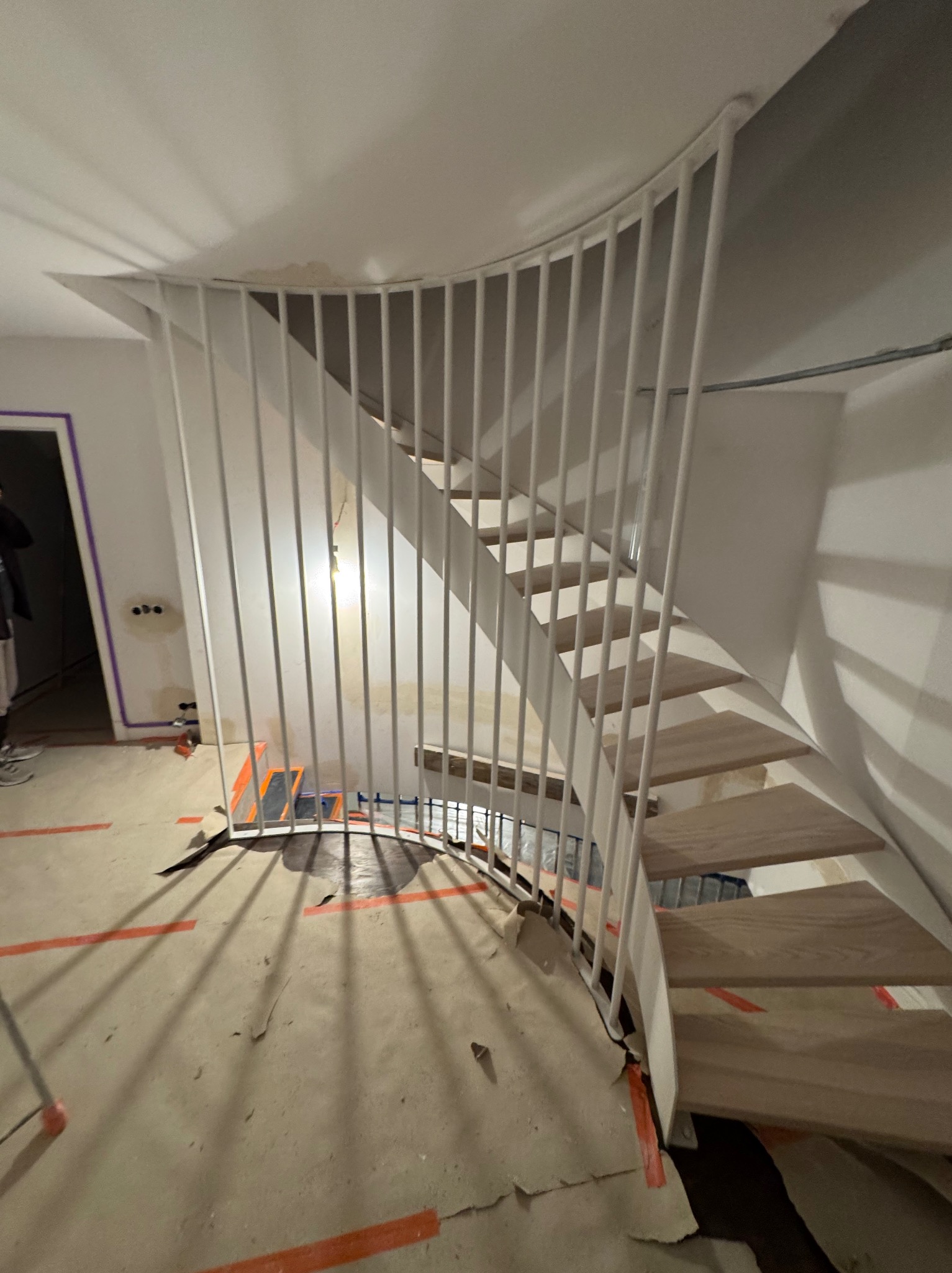 Spiralne schody z drewnianymi stopniami i białą, metalową balustradą w trakcie montażu. Widoczne zabezpieczenia podłogi i niedokończone wykończenie ścian.