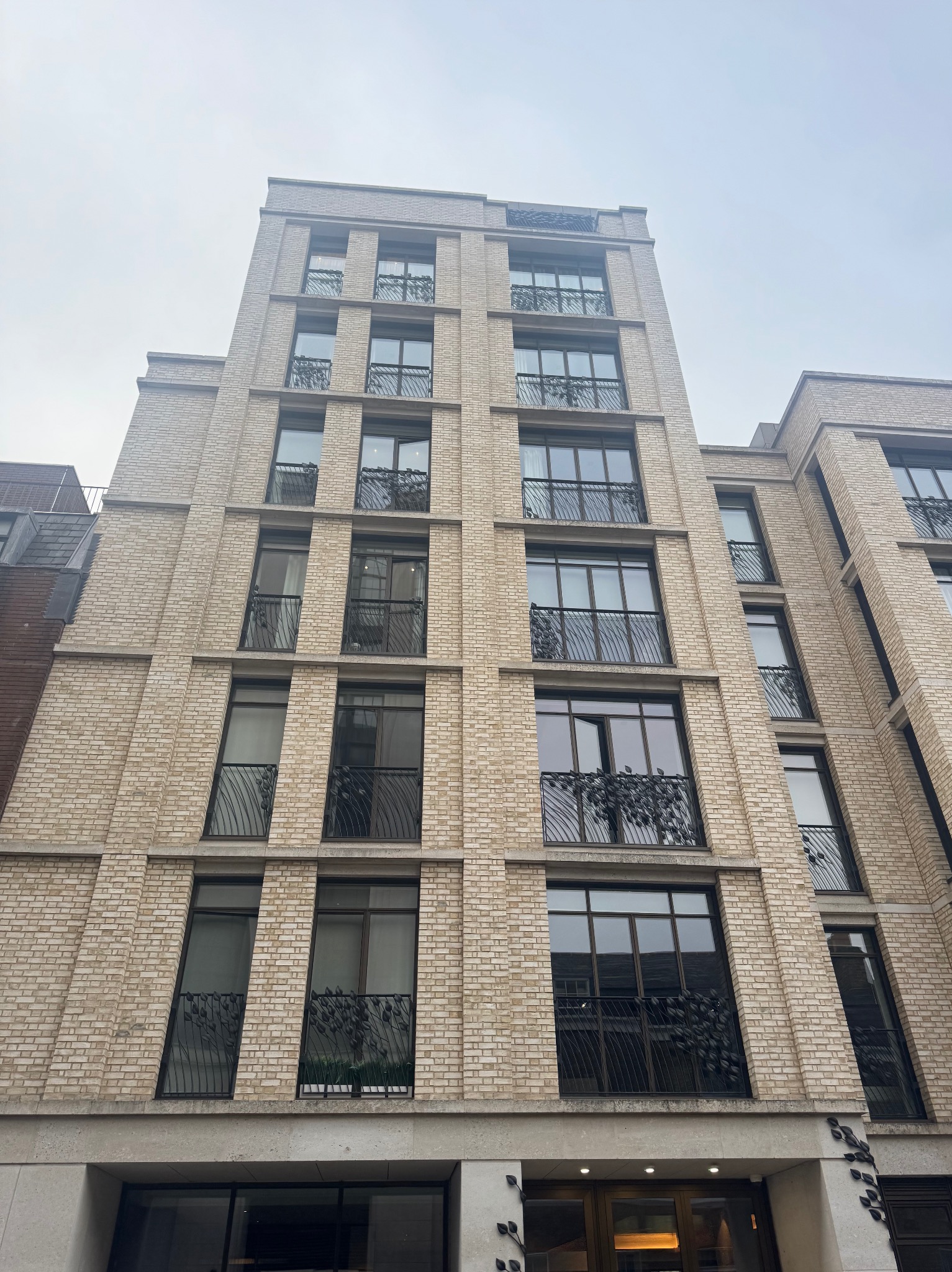 Elewacja budynku z jasnej cegły z balkonami z dekoracyjnymi, ciemnymi balustradami. Ujęcie z dołu podkreśla wysokość budynku i detale architektoniczne.