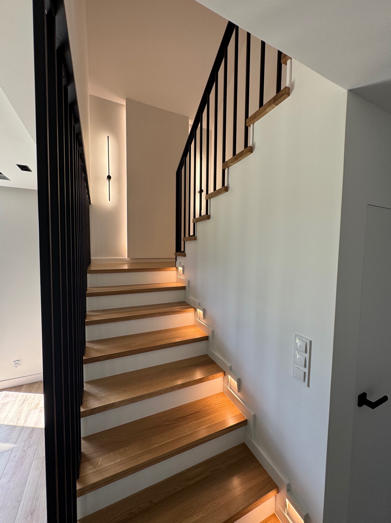 Nowoczesne schody drewniane z czarną, metalową balustradą i subtelnym oświetleniem LED wzdłuż stopni, w minimalistycznym wnętrzu z białymi ścianami.