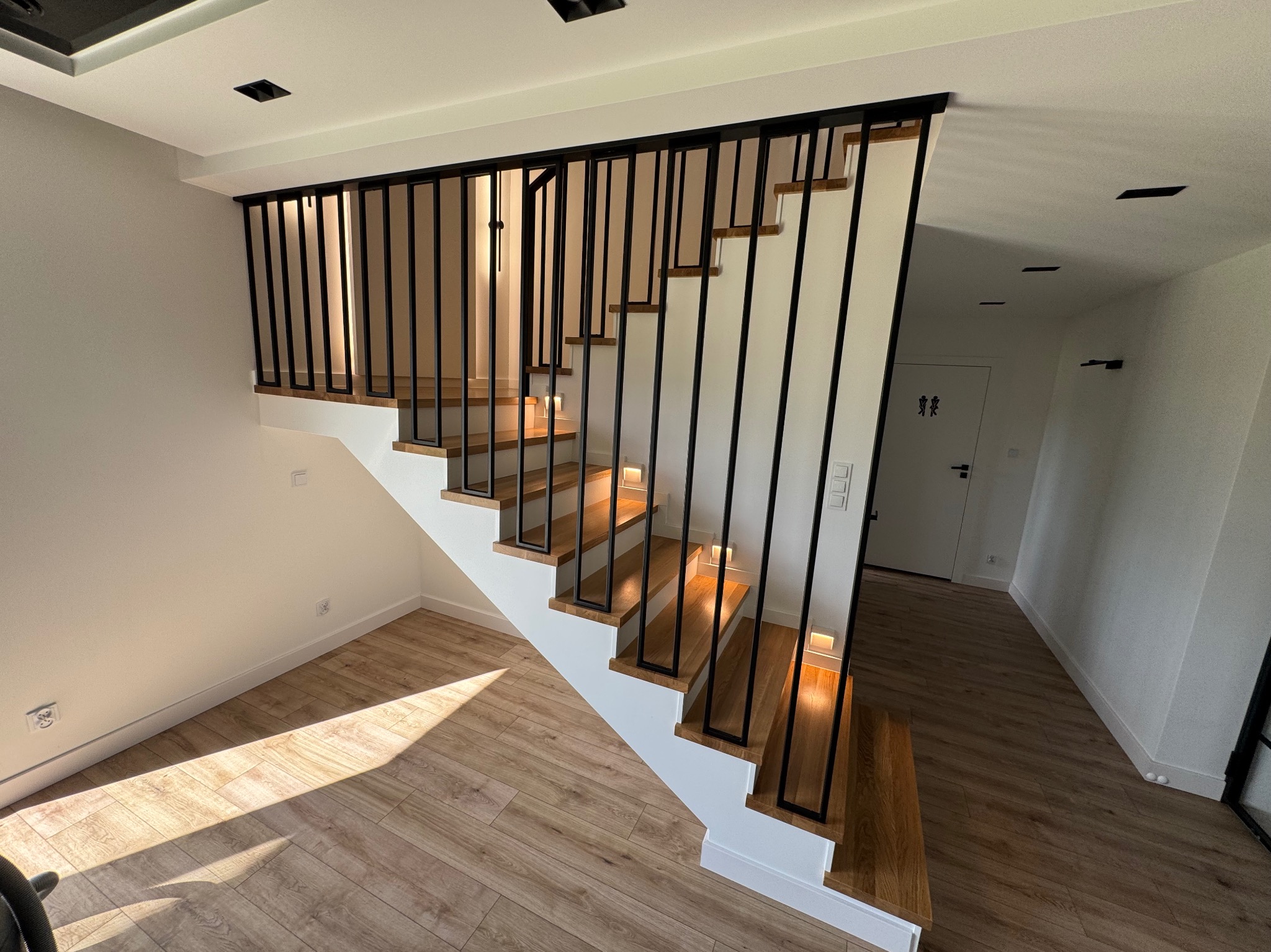 Nowoczesne schody drewniane z czarną, minimalistyczną barierką w jasnym wnętrzu. Widoczne oświetlenie schodów i drewniana podłoga. Ujęcie z dołu, podkreślające geometrię.