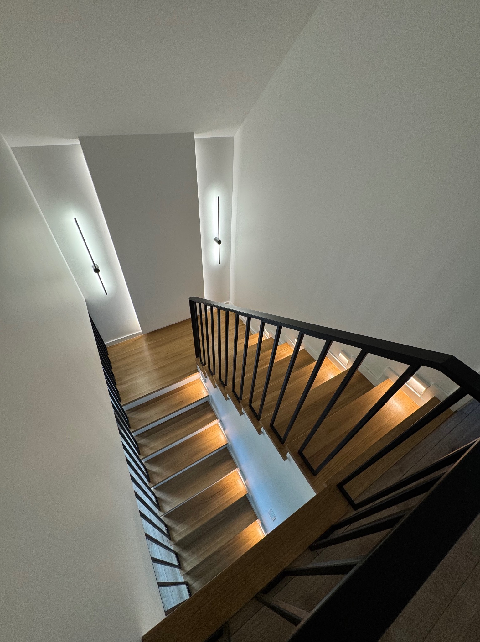 Nowoczesne schody drewniane z czarną balustradą, oświetlone punktowo, widziane z góry. Minimalistyczny design, białe ściany z podłużnymi lampami.