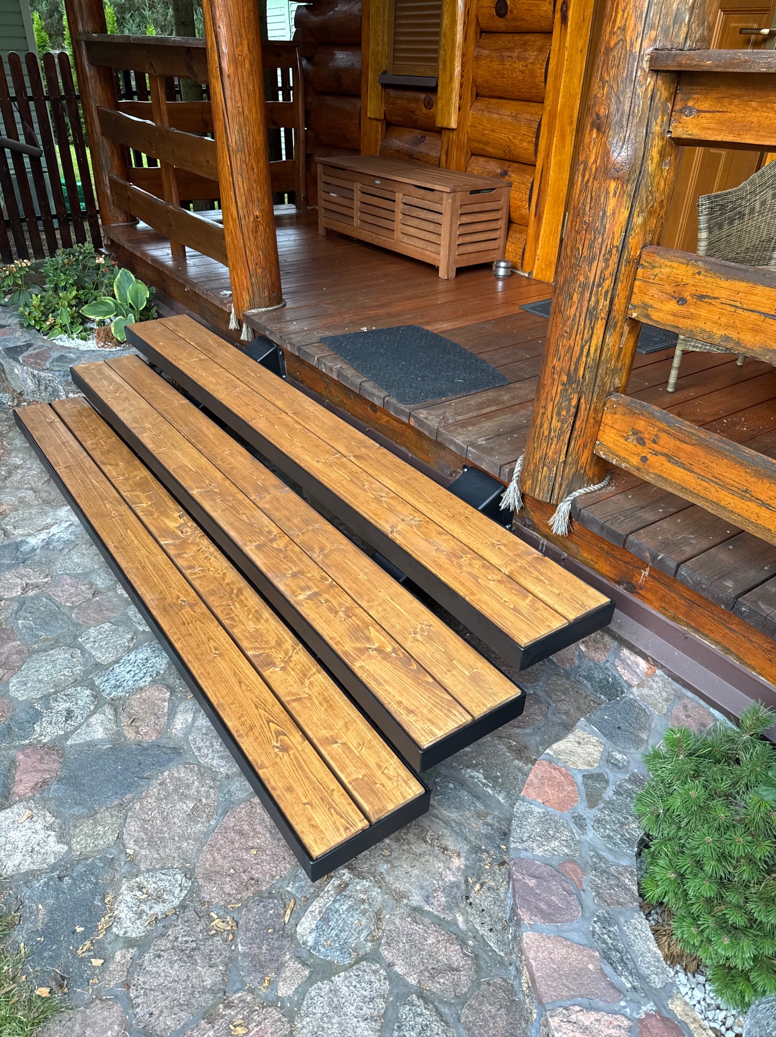 Drewniane schody wejściowe z czarną ramą, prowadzące do drewnianego domu z bali. Kamienna ścieżka i roślinność w tle. Ujęcie z niskiej perspektywy.