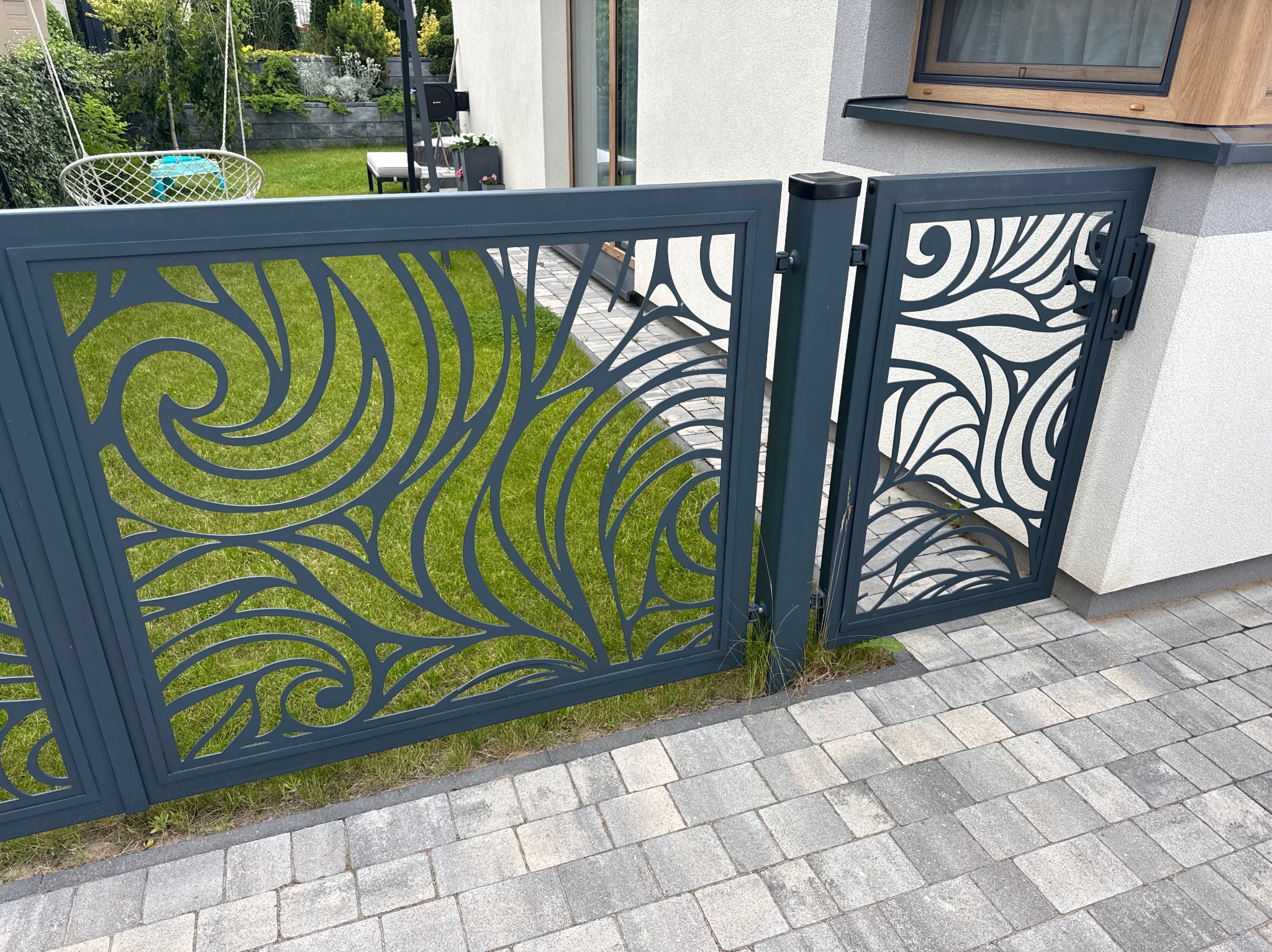 Nowoczesna, metalowa brama z ażurowym, roślinnym wzorem, pomalowana na grafitowo. Brama zamontowana przy jasnym budynku, widoczny trawnik i kostka brukowa.