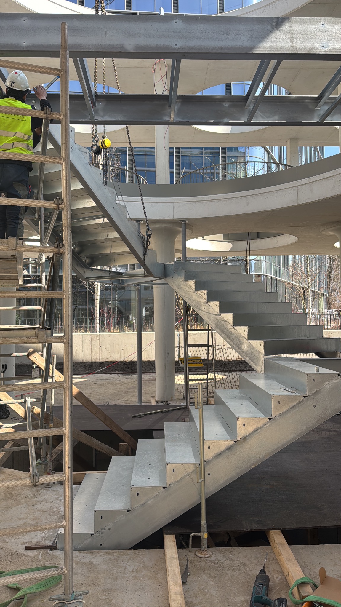 Montaż spiralnych schodów z metalu na zewnątrz budynku. Widoczny pracownik na rusztowaniu i elementy konstrukcji. Jasne światło dzienne.