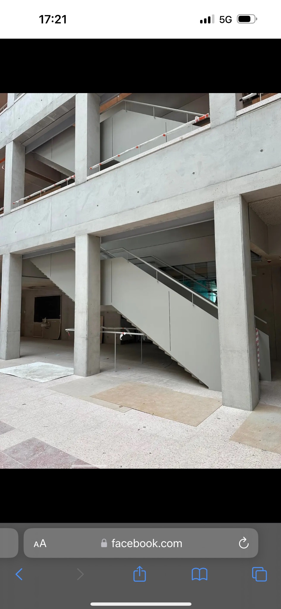 Wnętrze surowego budynku w trakcie budowy. Widoczne betonowe kolumny, strop i schody z jasną balustradą. Podłoga wyłożona płytkami. Ujęcie z dołu, podkreślające konstrukcję.