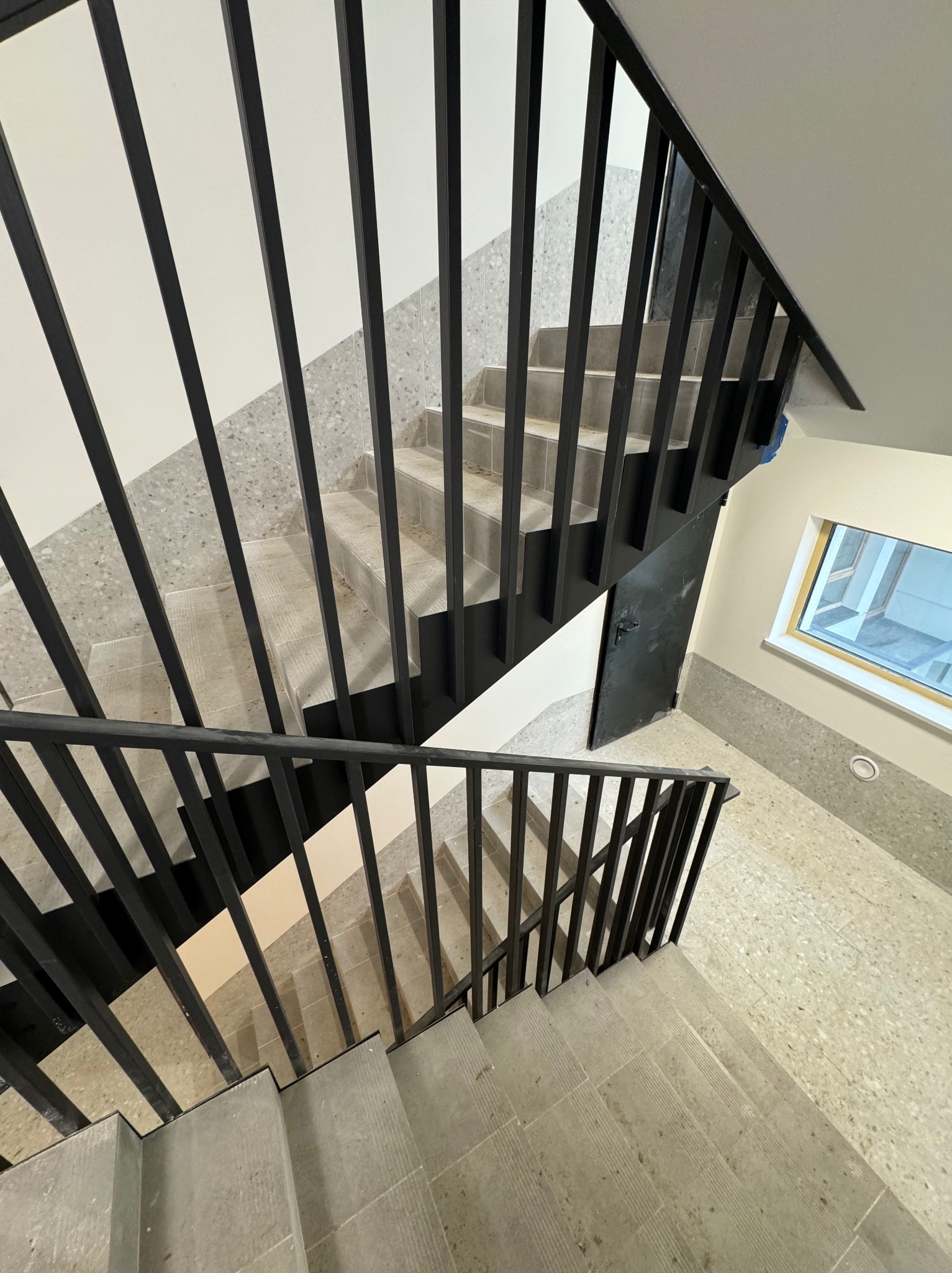 Metalowe schody z czarną, minimalistyczną balustradą, widok z góry. Szare płytki na schodach i podłodze, jasne ściany. Nowoczesne wykończenie.