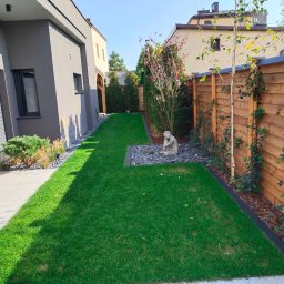 ,, KRAWCZYK '' F. U. H. - Zadbany trawnik z rzadko spotykaną rzeźbą, graniczący z drewnianym płotem, na którym pną się rośliny. Obok nowoczesny budynek z szarym wykończeniem.