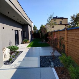 ,, KRAWCZYK '' F. U. H. - Nowoczesny ogród z minimalistycznym designem: jasne płyty chodnikowe, ciemne kamienie, drewniany płot i zadbana zieleń.