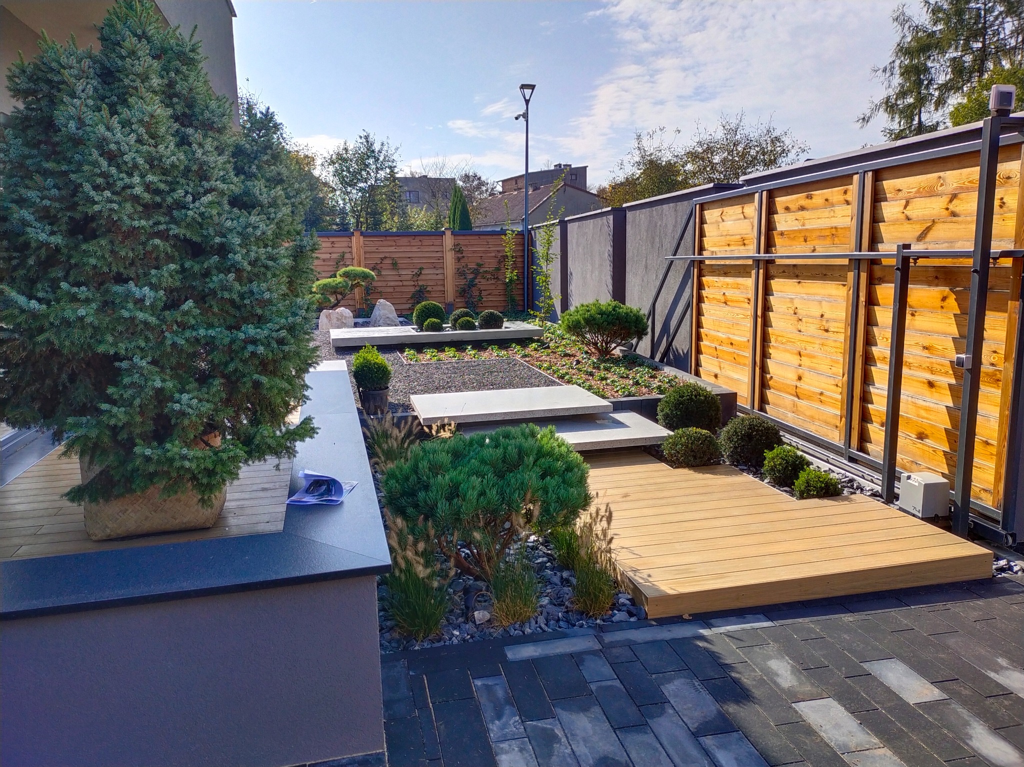 Nowoczesny ogród z drewnianym tarasem, ścieżką z płyt, ozdobnymi krzewami i kamieniami. Widoczny element drewnianego ogrodzenia i oświetlenie.