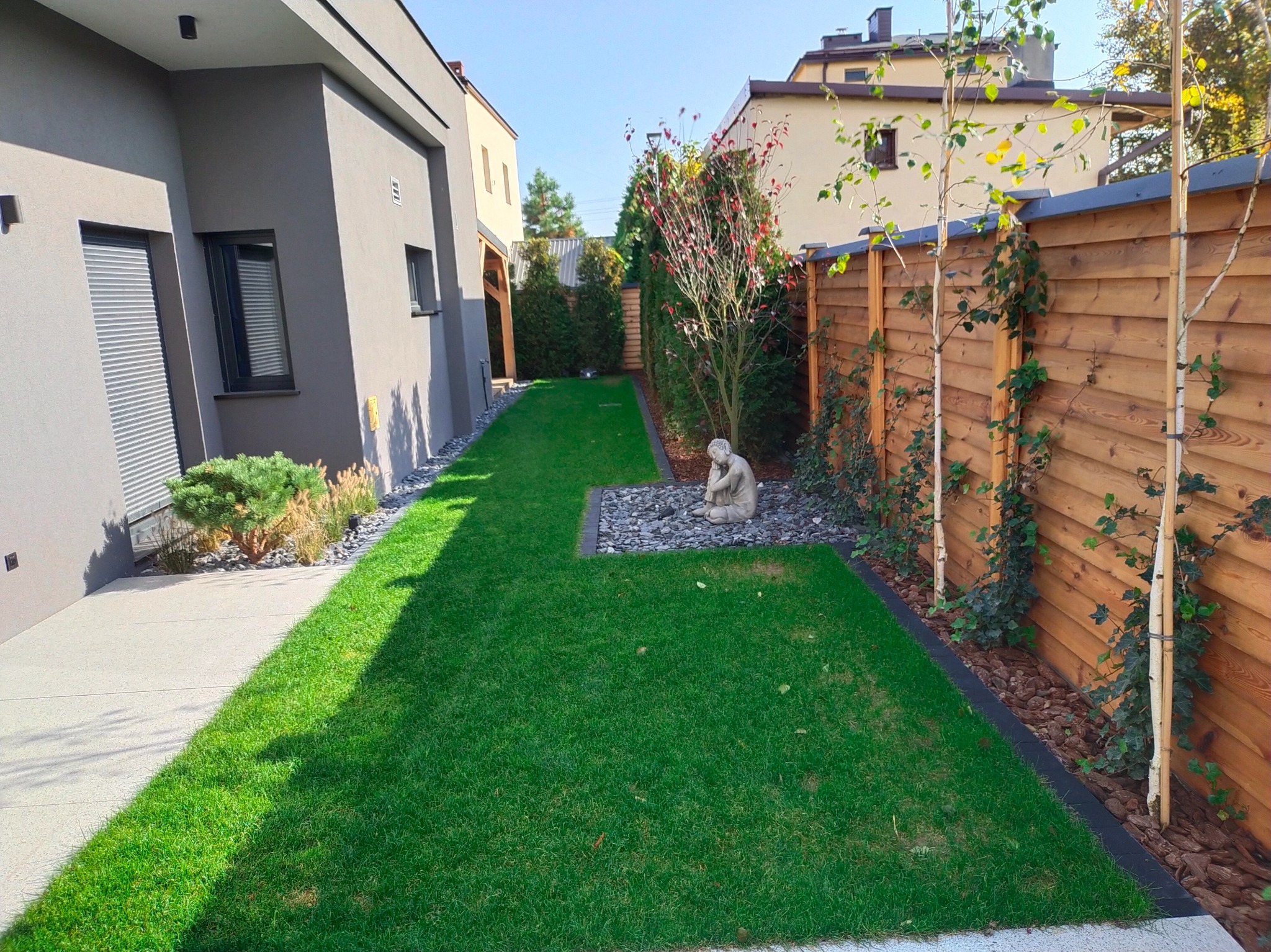 Zadbany trawnik z rzadko spotykaną rzeźbą, graniczący z drewnianym płotem, na którym pną się rośliny. Obok nowoczesny budynek z szarym wykończeniem.