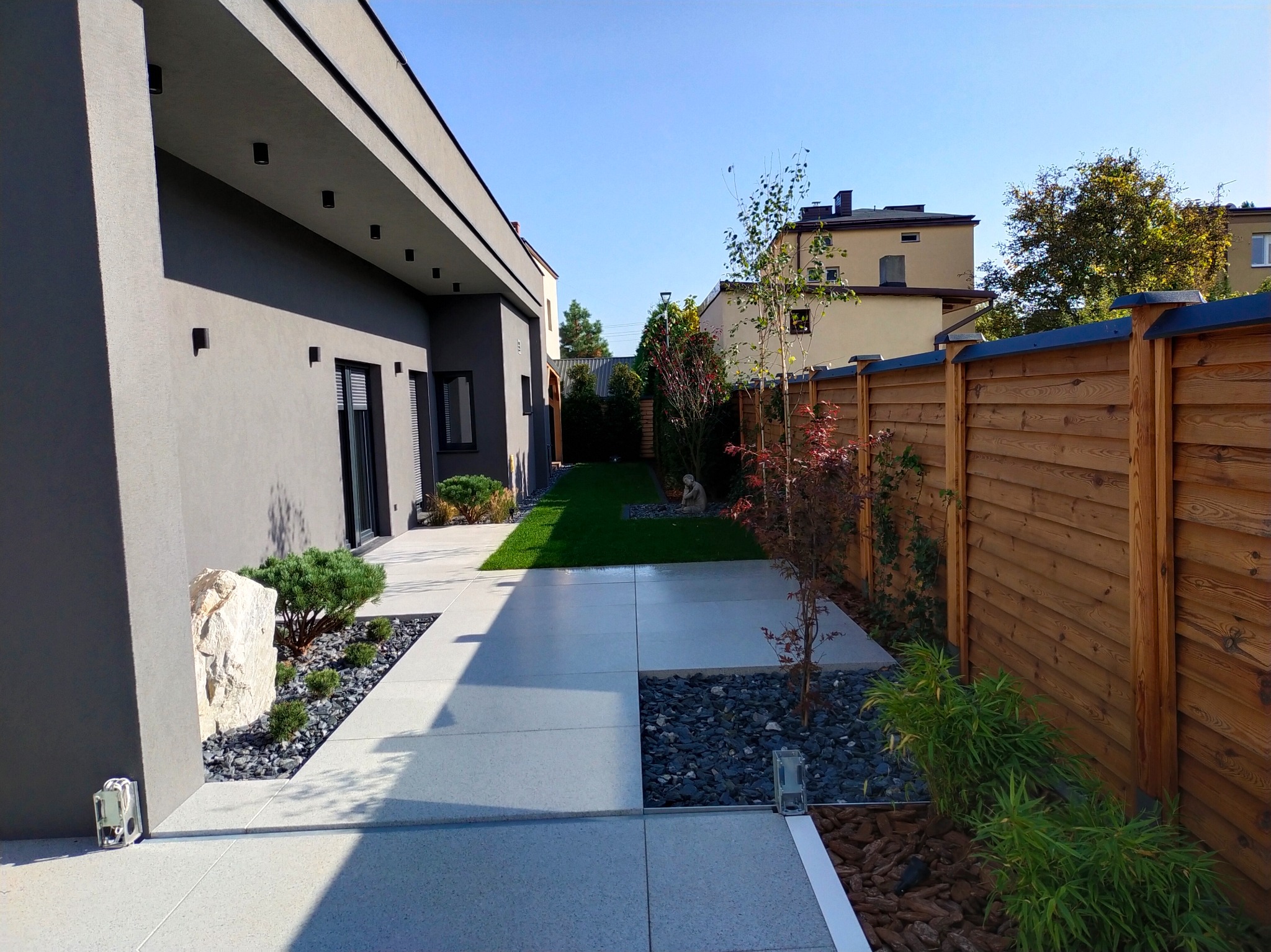 Nowoczesny ogród z minimalistycznym designem: jasne płyty chodnikowe, ciemne kamienie, drewniany płot i zadbana zieleń.