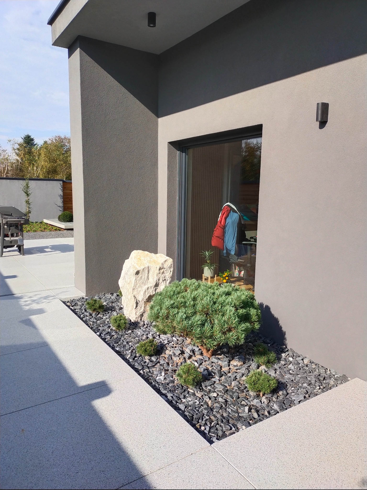 Nowoczesny ogród z rabatą przy budynku: kamień, karłowate sosny na czarnym żwirze. W tle widoczny grill i zadbany trawnik. Minimalistyczny design.