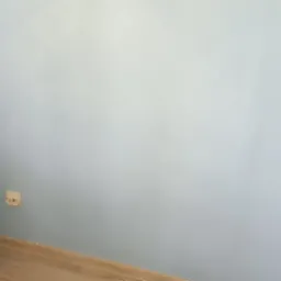 Ściana pomalowana na jasny odcień niebieskiego, widoczne dwa gniazdka elektryczne, fragment drewnianej podłogi i zasłony w lewym rogu.