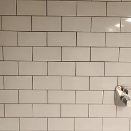 Białe, prostokątne płytki ułożone w cegiełkę na ścianie, z widoczną chromowaną baterią wannową. Minimalistyczny design łazienki.