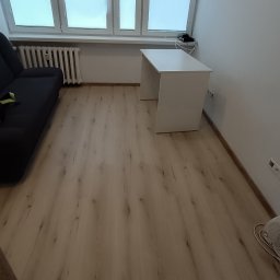 Centrum Edukacji Focus Jakub Mańczyński - Jasne panele podłogowe w pokoju z szarą sofą i białym biurkiem pod oknem. Widoczne listwy przypodłogowe i grzejnik. Minimalistyczne wnętrze po remoncie.