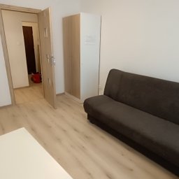 Jasne wnętrze pokoju po remoncie: beżowe panele podłogowe, szara sofa, biała szafa i drzwi. W rogu widoczne materiały budowlane, sugerujące świeżo zakończone prace.