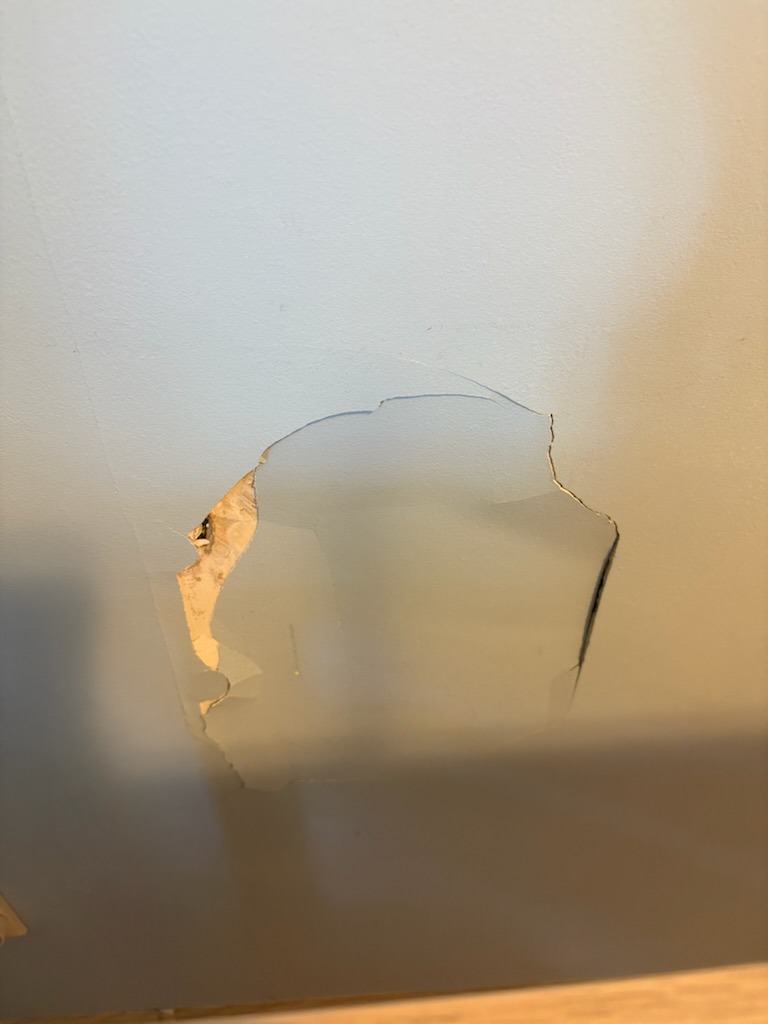 Uszkodzona ściana z widocznym ubytkiem tynku i farby, ukazująca warstwy podkładowe. Fragment drewnianej listwy przypodłogowej u dołu kadru.